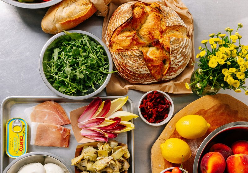 An overhead spread of fresh bread, tomatoes, mozzarella, prosciutto, and produce on a kitchen counter